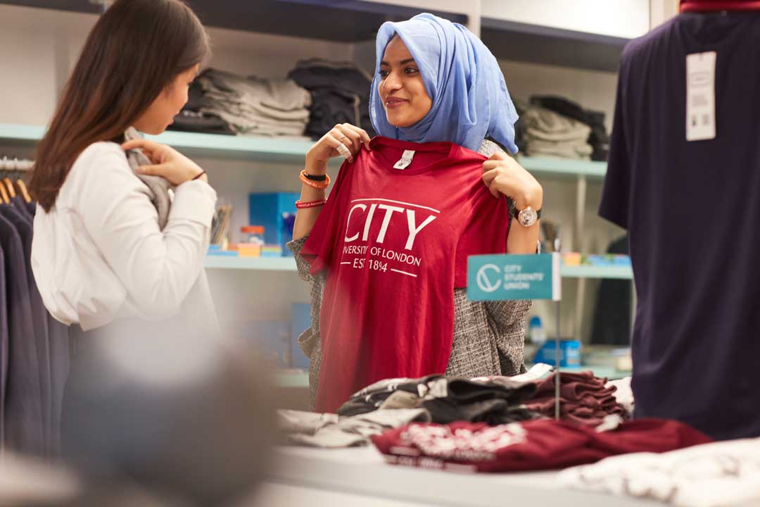 A student wearing a hijab holding up a City SU t-shirt against their body to show a friend